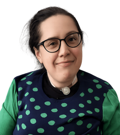 A woman with glasses and a blue and green spotted top smiles at the camera in front of a plain background.