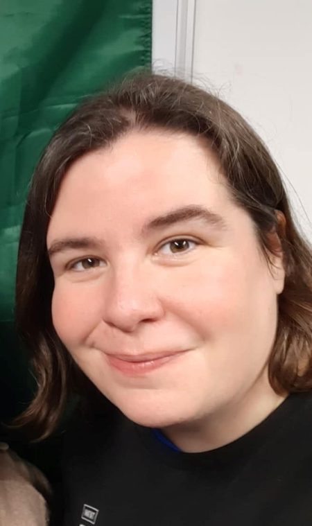 A person with short brown hair smiles whilst wearing a black shirt, standing before a green and white background.