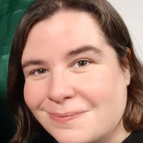 A person with short brown hair smiles whilst wearing a black shirt, standing before a green and white background.