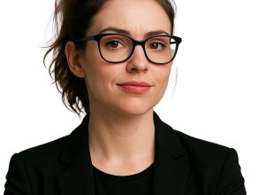 A woman with a brown ponytail, black glasses, and a black blazer stands in front of a white background.