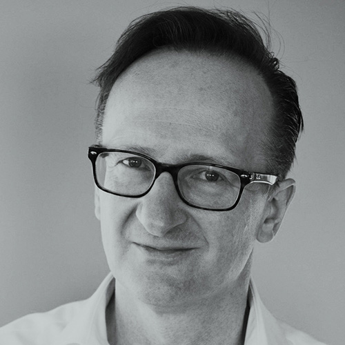 A man with short hair and dark sunglasses, dressed in a collared shirt, smiles slightly in a black and white photograph.