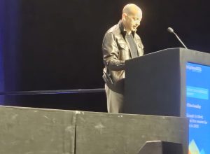 A man wearing a black jacket speaks into a microphone at a lectern on a dimly lit stage.