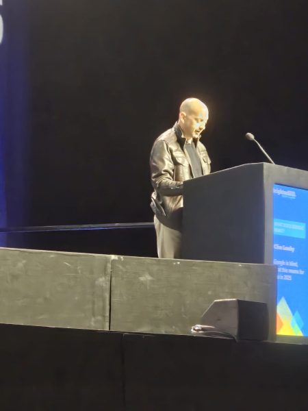 A man wearing a black jacket speaks into a microphone at a lectern on a dimly lit stage.