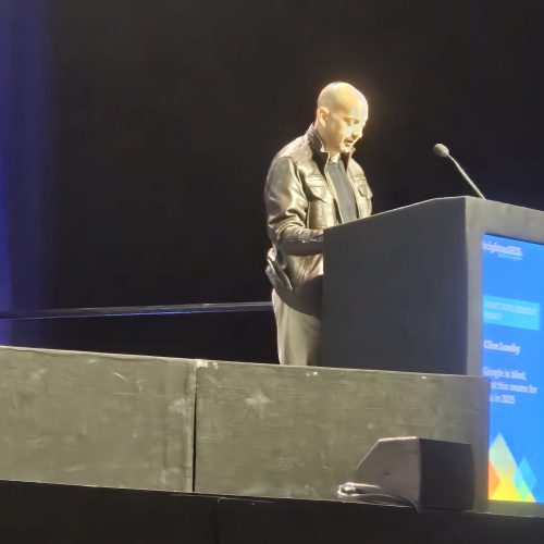 A man wearing a black jacket speaks into a microphone at a lectern on a dimly lit stage.