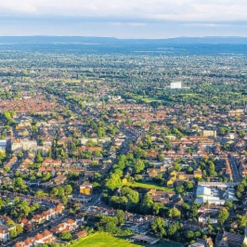 Aerial photo of a city featuring grid-patterned streets, residential houses, patches of green space, and distant rolling hills.