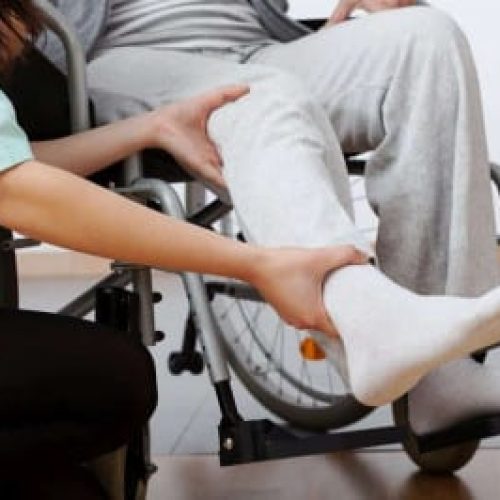 A carer assists a wheelchair user by supporting their leg, aiding in movement or therapeutic exercise.