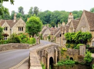 A stone bridge crosses towards a village of old stone cottages surrounded by lush green trees and plants.