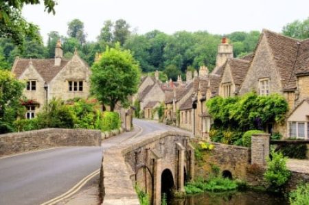A stone bridge crosses towards a village of old stone cottages surrounded by lush green trees and plants.