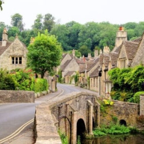 A stone bridge crosses towards a village of old stone cottages surrounded by lush green trees and plants.