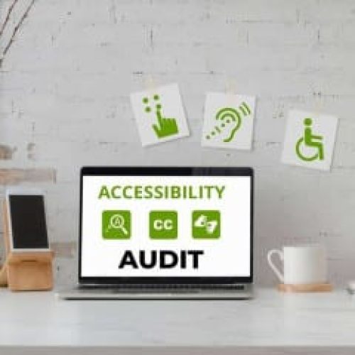 A laptop sits on a desk showing Accessibility Audit and related icons, with decor and accessibility signs visible on the wall behind.