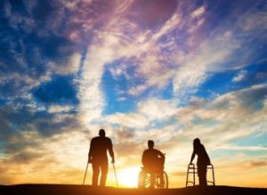 Three individuals using mobility aids—crutches, a wheelchair, and a Zimmer frame—are silhouetted against a vibrant sunset sky.