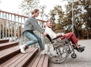 A woman helps a man in a wheelchair navigate down a ramp next to an outdoor stairway.