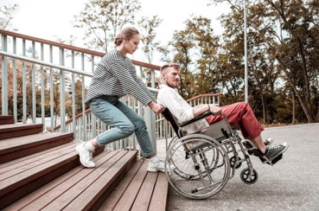 A woman helps a man in a wheelchair navigate down a ramp next to an outdoor stairway.