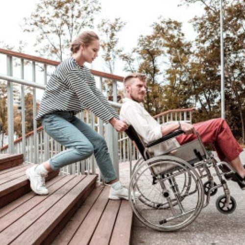 A woman helps a man in a wheelchair navigate down a ramp next to an outdoor stairway.