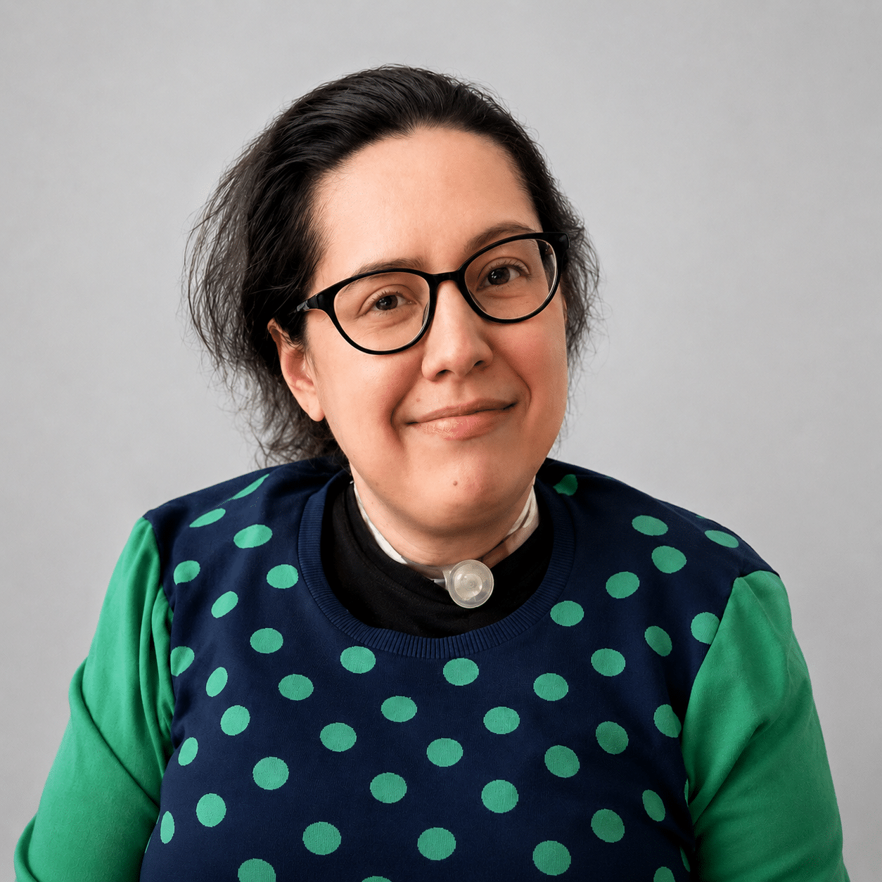 A smiling person with glasses and dark hair, dressed in a blue shirt with green polka dots and green sleeves, sits at a computer working on website accessibility for a test web project.