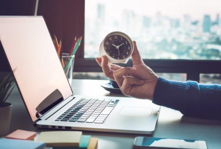 A person holds a small clock next to a laptop on a desk, with a cityscape visible through the window behind them.