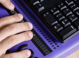A person’s hands use a braille display on a purple desk with a computer keyboard in the background.