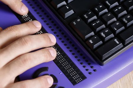 A person’s hands use a braille display on a purple desk with a computer keyboard in the background.