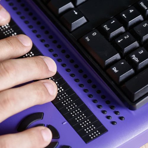 A person’s hands use a braille display on a purple desk with a computer keyboard in the background.