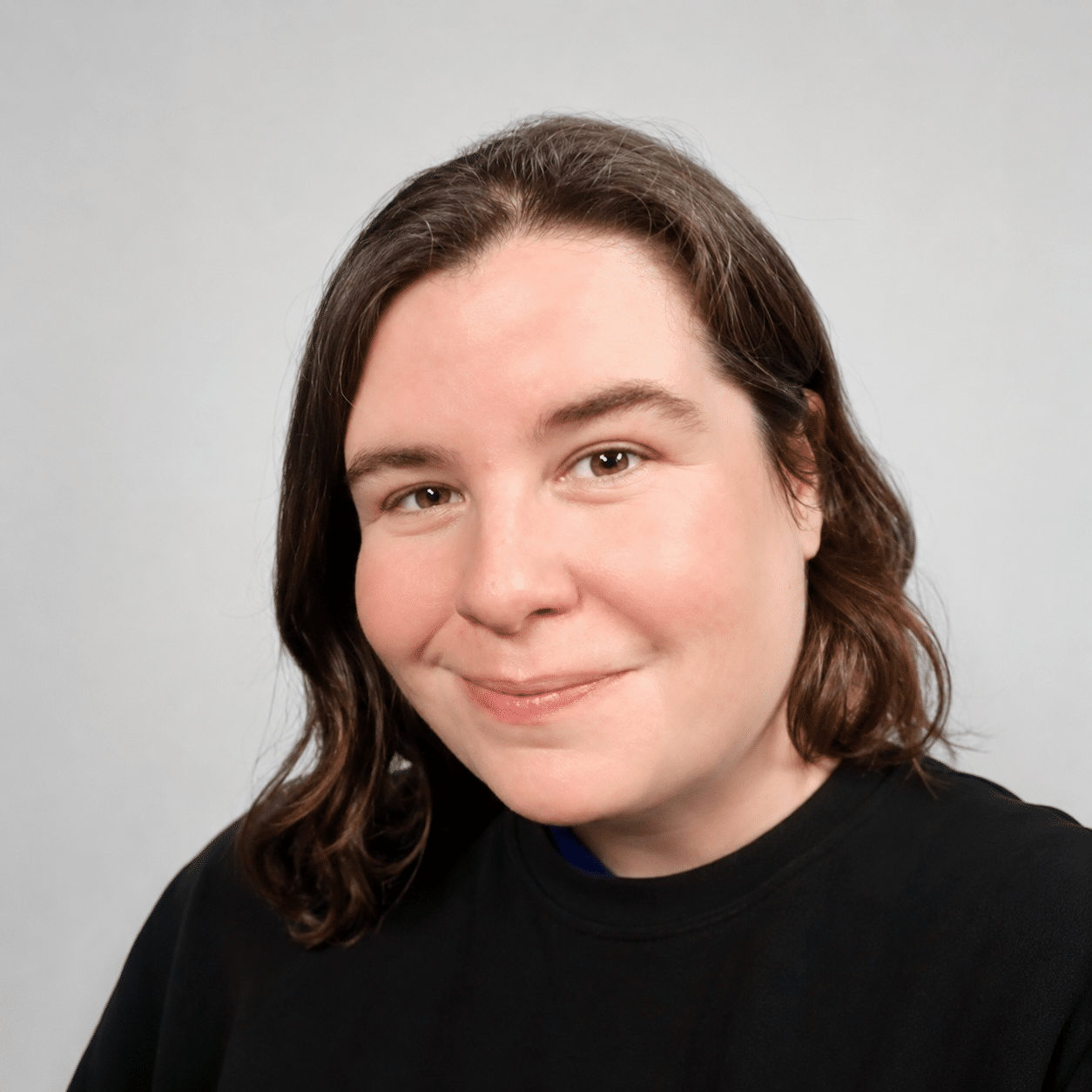 A person with medium-length brown hair is smiling and wearing a black shirt, standing in front of a plain light background.