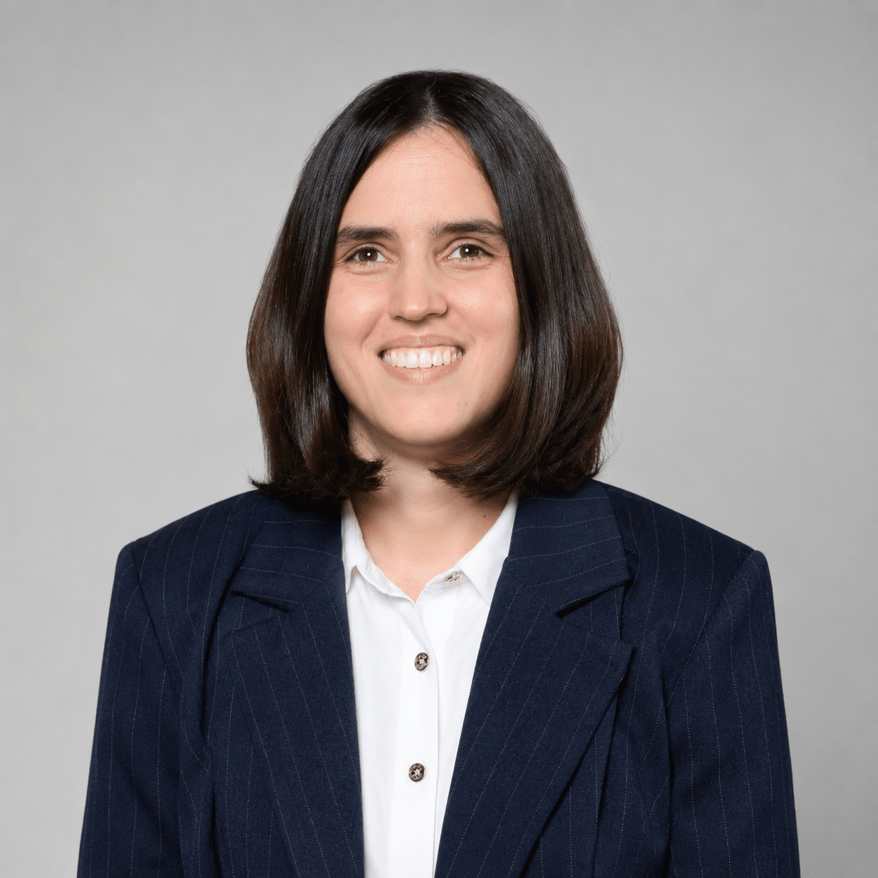 Woman wearing a navy pinstripe blazer and white shirt, smiling in front of a plain light grey background.