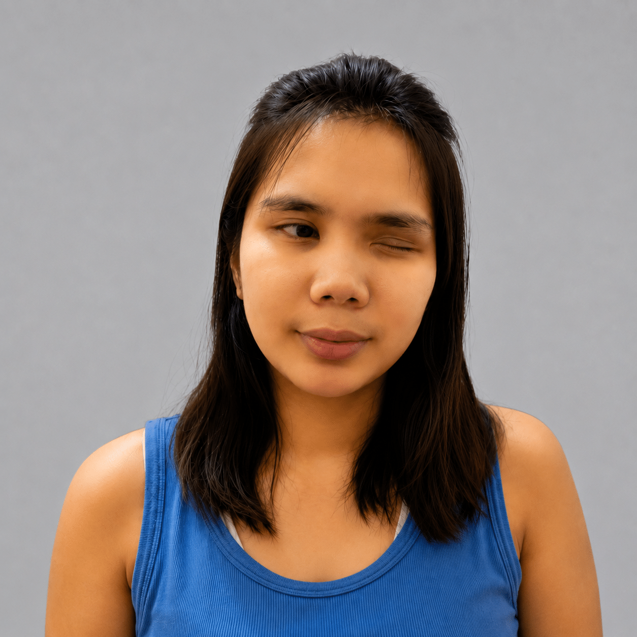 A woman in a blue vest winks with one eye, standing against a plain grey background, appearing ready to test web design concepts or check website accessibility.