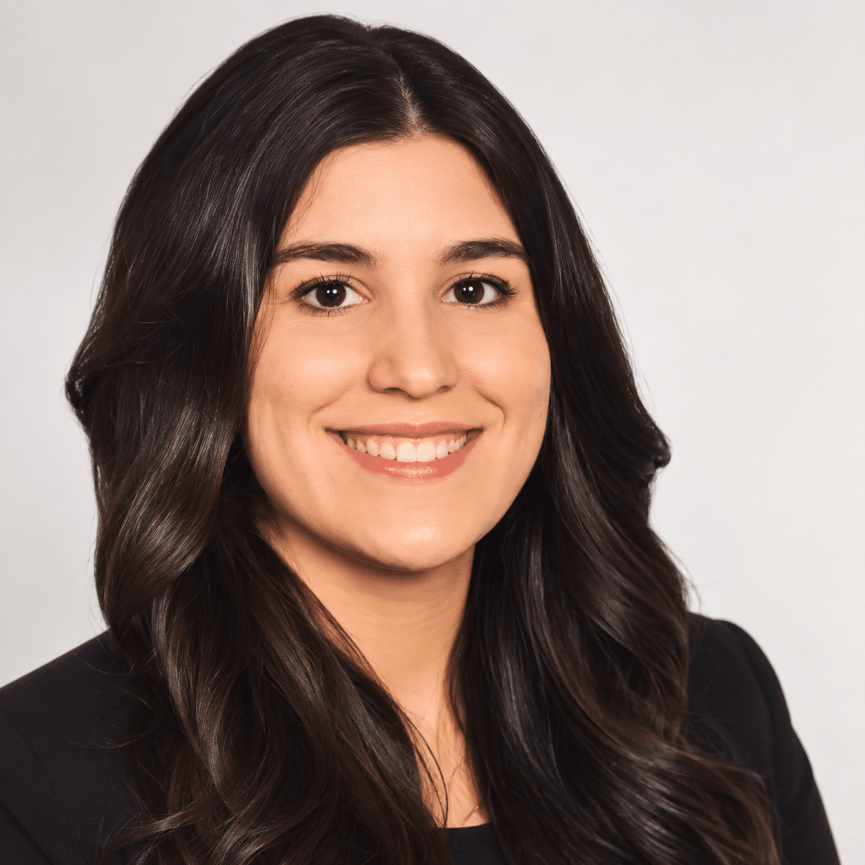 A smiling woman with long dark hair wears a black blazer and stands against a plain light background, portraying confidence and professionalism.