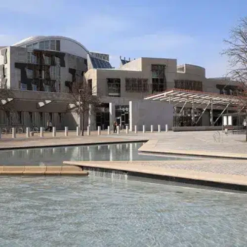 A modern building with distinct architecture stands behind a shallow pool and several leafless trees beneath a blue sky.