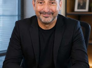 A bald man wearing a black jacket sits at a desk, smiling, with shelves and a photo frame behind him as he gets ready to check website accessibility.
