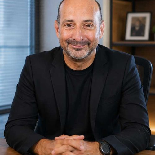 A bald man wearing a black jacket sits at a desk, smiling, with shelves and a photo frame behind him as he gets ready to check website accessibility.