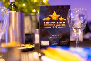 A promotional display for the Chichester & Bognor Business Awards 2024, featuring the event program on a table set with wine glasses and cutlery.