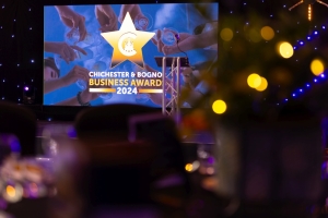 A stage with a screen displaying Chichester+ Bognor Business Award 2024 featuring a gold star and crescent moon logo. The foreground is softly blurred, with hint of decorated tables and lights, indicating a celebratory atmosphere.