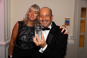 Chichester-Bognor-Business-Awards-2024-29-wClive and his partner Sarah are in formal attire and are smiling at the camera, with the Clive proudly holding a clear glass award. Sarah has blonde hair and is wearing a black dress, while the Clive is bald with a bow tie and tuxedo.