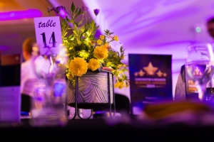 A table decorated with a potted plant with yellow flowers, a "table 14" sign, and award plaques in the background. The ambiance is set with purple lighting. The focus is on the floral arrangement with bokeh effects in the background.