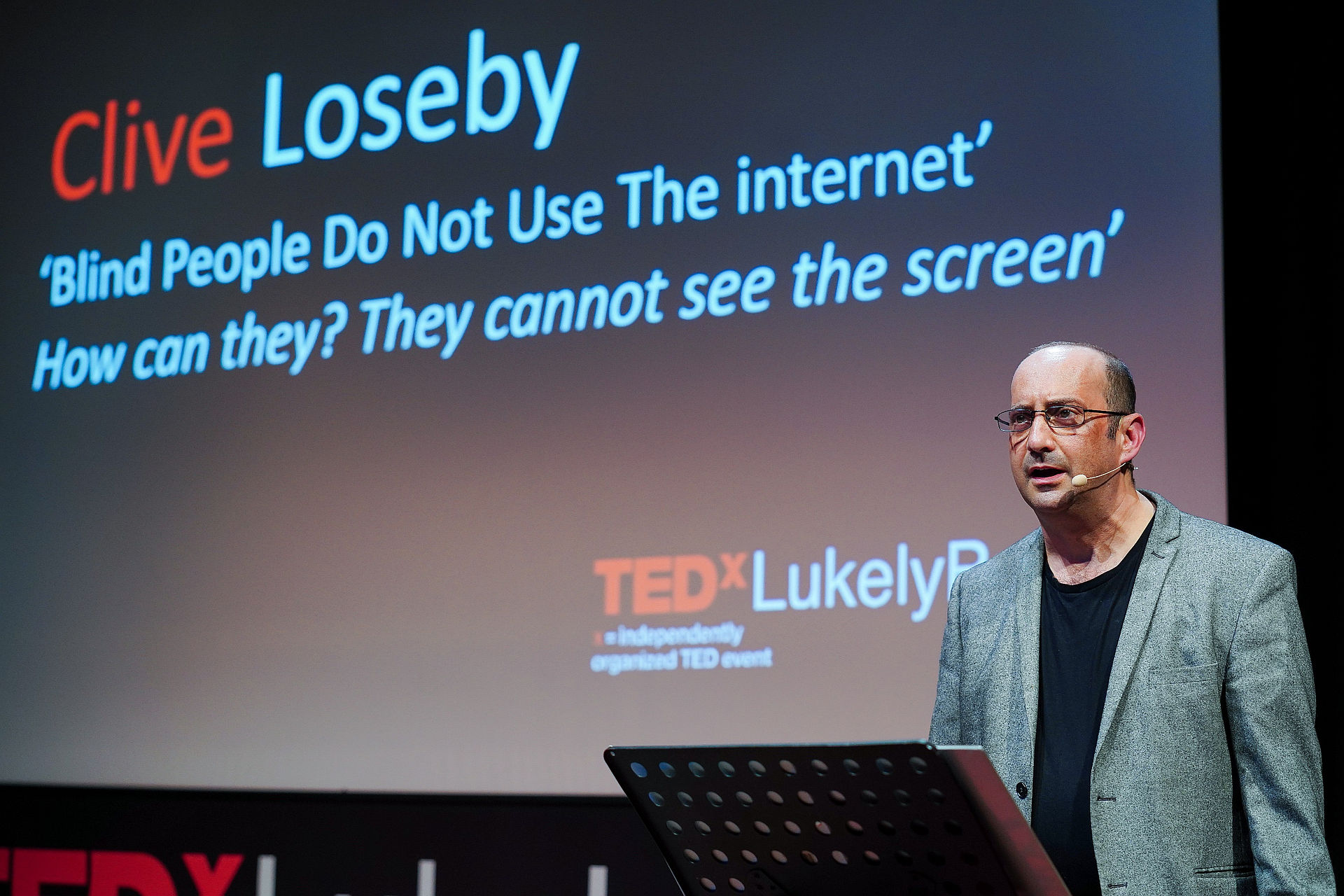 Clive giving his Ted Talk. Behind him, a screen says "Blind People do not use the Internet. How can they? They cannot see the screen"