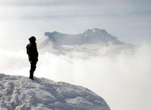 Man on a mountain is gazing into the distance, across a misty landscape. A snow-covered mountain range is far off.