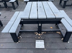 A table with 2 benches fixed to it, gaps are on the other sides for wheelchairs. A sign on the floor reads: Polite Notice Please prioritise these tables for disabled users