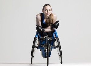 a young female athlete is sitting in a racing wheelchair. She has long hair, a pony tail, blue eyes, no legs and is wearing black leather gloves.