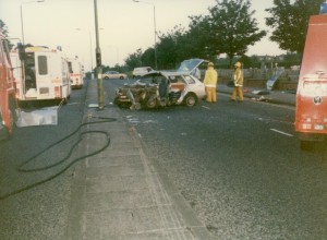 A traffic accident scene showing a significantly damaged car in the middle of the road. Firefighters in yellow gear are on-site, with emergency vehicles including fire engines and an ambulance present. Hoses are laid out on the ground, and trees line the street.