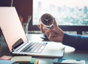 A hand holding a small clock is extended over a laptop on a desk. The desk also has colorful sticky notes, pencils, and a blurry cityscape is visible through the window in the background.