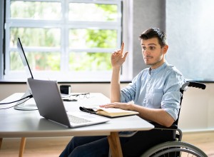 A man sitting in a wheelchair at a desk with a laptop, raising his right hand. He appears to be asking a question or participating in an online meeting. An open notebook and eyeglasses are on the desk. The background shows large windows with greenery outside.