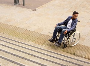 A person in a wheelchair, dressed in a blue blazer and jeans, looks to their right at the base of a flight of stairs. They are on a paved outdoor area with a lamppost in the background.