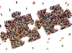 A large group of people is gathered to form two interlocking puzzle pieces against a white background. Additional smaller groups and individuals are scattered around the central formation, creating a sense of movement and interaction among the crowd.