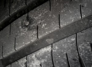 A close-up image of a worn car tire with visible cracks and a nail embedded in the tread. The tire\'s surface shows signs of wear and tear, indicating it has been used extensively and requires repair or replacement.