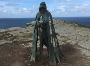 A bronze statue of a cloaked figure stands on a rocky cliffside with the ocean in the background. The cloak appears tattered and the figure holds a staff. The statue is set against a backdrop of a partly cloudy sky and rugged terrain.