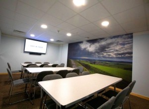 A conference room with white rectangular tables and gray chairs in front of a mural depicting a landscape with green fields and a cloudy sky. There is a wall-mounted screen displaying the text ONE O FOUR. The room has a modern ceiling with recessed lighting.