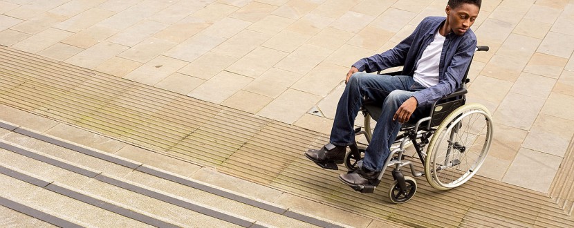 A person in a wheelchair, dressed in a blue blazer and jeans, looks to their right at the base of a flight of stairs. They are on a paved outdoor area with a lamppost in the background.