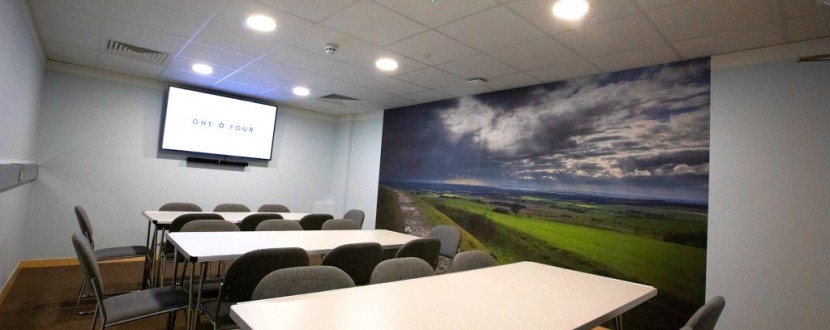 A conference room with white rectangular tables and gray chairs in front of a mural depicting a landscape with green fields and a cloudy sky. There is a wall-mounted screen displaying the text ONE O FOUR. The room has a modern ceiling with recessed lighting.