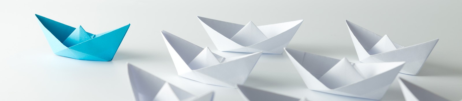 A group of white origami paper boats with one distinct blue origami paper boat standing out among them, arranged on a plain white surface.