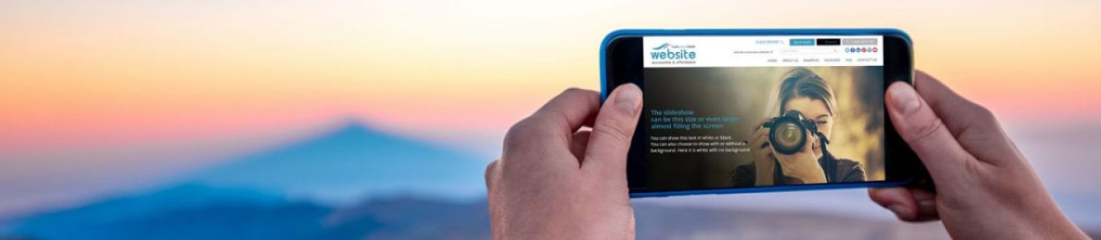 A person wearing a blue jacket holds a smartphone horizontally to take a photo of a scenic landscape at sunrise or sunset. The image on the phone's screen shows another person photographing the viewer. The background features rolling hills under a colorful sky.